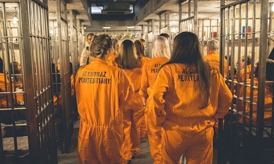 A group of 8 ladies in orange jumpsuits walking into cells.